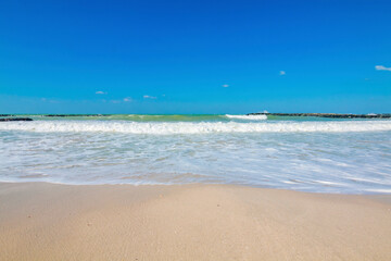Pristine beach in Sharjah on a sunny day, with golden sand stretching along the shoreline. The calm turquoise waters gently lap against the shore, under a clear, cloudless blue sky.