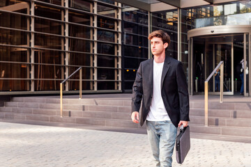 young adult man in casual suit walking in front of an office building holding a briefcase, concept...