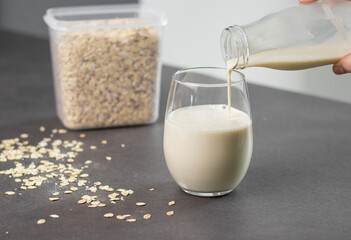 Close up of a female hand pouring oat milk into a glass on grey kitchen table. Vegan and non-dairy alternative milk