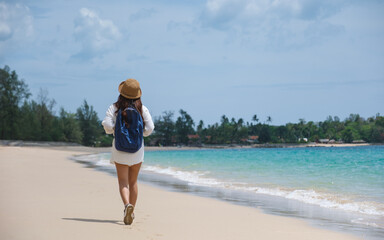 Rear view of a female traveler with hat and backpack standing on the beach by the sea