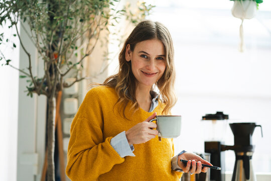 Woman in yellow sweater holding a cup of tea and smartphone indoors