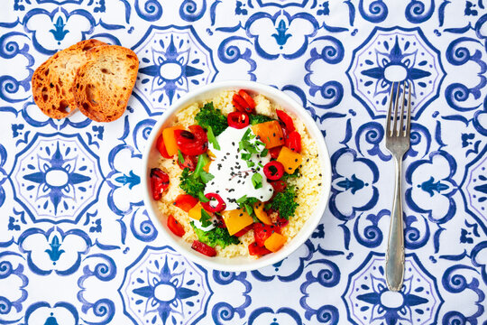 Bowl of couscous with grilled vegetables and yoghurt sauce on patterned tablecloth