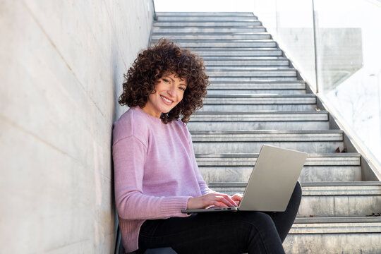 Woman with curly hair sitting on stairs working on a laptop and smiling outdoors