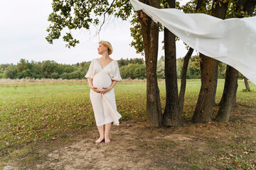 Pregnant woman in a white dress standing barefoot in nature, surrounded by trees, embodying serenity and natural motherhood.