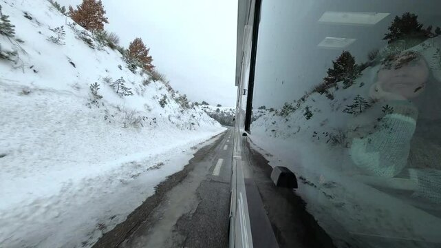 Woman seen through the camper window as she travels on a snowy road in winter, with the motorhome&rsquo;s exterior and a cold landscape featuring snow-covered roads and a gray sky