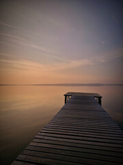 A serene view of a wooden dock extending into a vast body of calm water at sunset. the sky is...