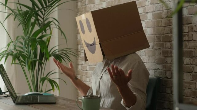 Unidentified office worker with cardboard box on head sitting at desk and using laptop, symbolizing online anonymity, restricted privacy, faceless corporations, and bureaucratic control