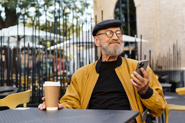 A senior man, in a trendy jacket and cap, sits at cafe table, sipping coffee and talking by phone.