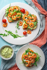Healthy shrimp with avocado and arugula on toast for breakfast on a white plate on a light blue background. View from above. Flat layout.