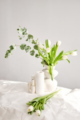 Composition of white tulips, eucalyptus and candles in a decorative vase. Light interior minimalism with spring mood, blurred foreground. Copy space. Still life