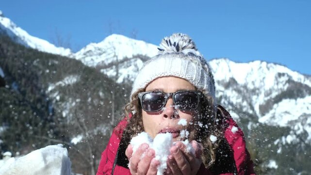 Young woman blowing a snowball in a snowy mountain during winter vacation; wearing a red jacket and colorful wool cap, she enjoys playful holiday moments in a crisp landscape beneath bright sky