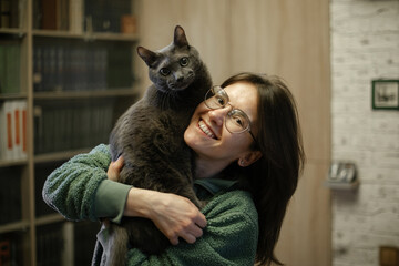 Young woman in glasses is happy and laughing holding a cat in her arms. In the background is a home interior.