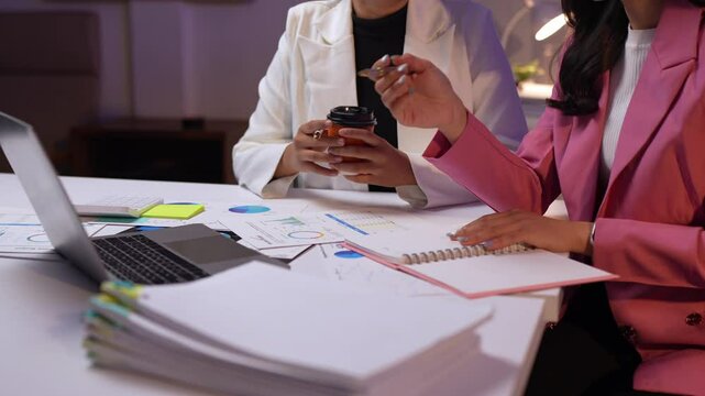Two businesswomen are working late in the office, analyzing financial reports on a laptop and taking notes, with a stack of documents and takeaway coffee cups on the table