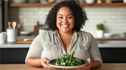 A woman holding a plate of greens stands in a kitchen with a warm, inviting ambiance, smiling towards the camera with pride or anticipation, suggesting she has prepared or made this dish herself.