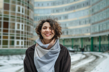 Smiling woman with curly hair wearing a scarf and coat in a snowy city setting