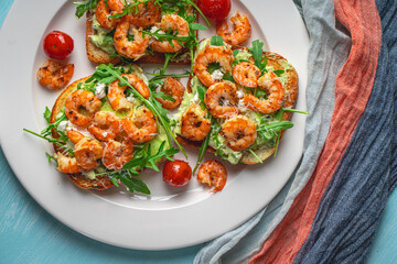 Toast or sandwiches with avocado oil, arugula and fried prawns on a white plate on a light blue background with a napkin. Healthy breakfast. View from above. Flat layout. Space for text.