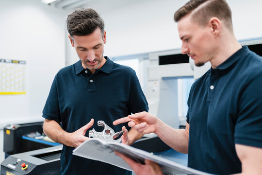 Male engineers with instruction manual examining machine part in factory office