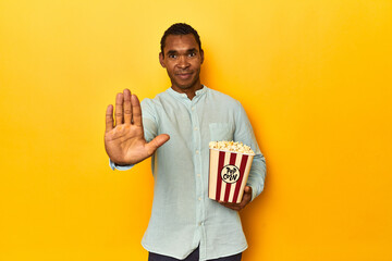 African American man with popcorn, yellow studio, standing with outstretched hand showing stop...