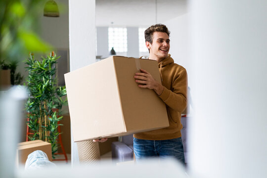 Smiling young man holding cardboard box while looking away in new apartment