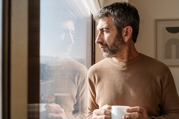 Man with gray hair looking out a window holding a cup indoors