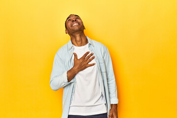 African American man in blue shirt, yellow studio, laughs out loudly keeping hand on chest.