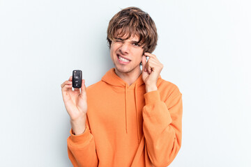 Young caucasian man holding car keys isolated on blue background covering ears with hands.