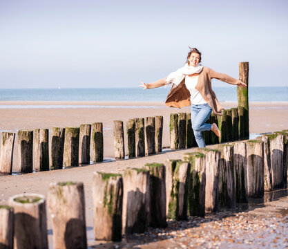 Smiling young woman with arms outstretched running amidst wooden posts at beach during sunny day