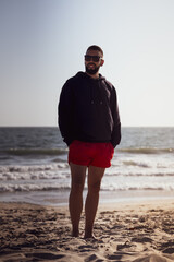Young man with beard and glasses on the shore of the beach in Santa Monica, California wearing a sweatshirt and swimsuit.