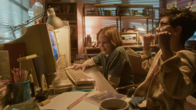 Two smiling young boys playing videogames using retro computer together in nostalgic 90s setting, with friend winning and cheering