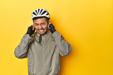 Asian man in bike helmet and gloves, yellow studio backdrop covering ears with hands.