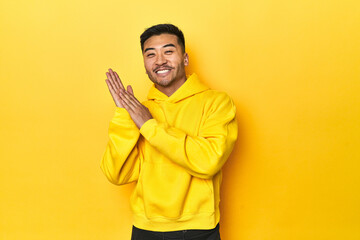 Bold Asian man in yellow hoodie on yellow studio feeling energetic and comfortable, rubbing hands confident.