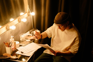 Person sitting in dressing room reading script and concentrating before performance with makeup items and desk lighting visible