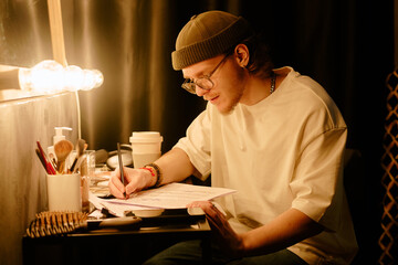 Young man focused on studying and writing notes at desk under warm lamp light, wearing beanie and glasses. Surrounded by study materials and personal items