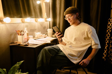 Individual sitting in front of mirror and using smartphone in well-lit room. Various items and...