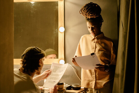 Team of filmmakers reviewing script in backstage area with thoughtful expressions, preparing for upcoming scenes. Lighting coming from surrounding lamps illuminating faces and workspace