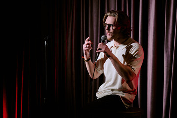 Comedian holding microphone while performing stand-up comedy routine on stage under spotlight with...