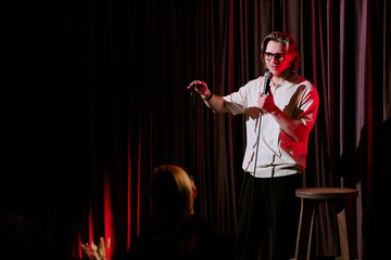 Comedian performing stand-up routine on stage, holding microphone and engaging with audience under...