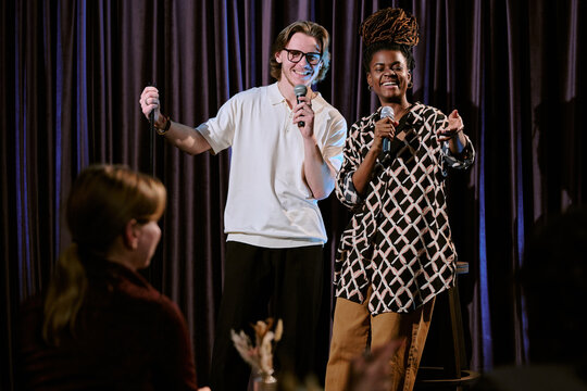 Man and woman comedians performing stand-up comedy on stage holding microphones, engaging audience with their jokes and energy
