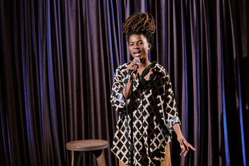 Female comedian performing stand-up comedy on stage holding microphone with stool in background....