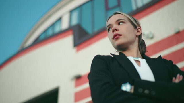 Young damsel in black blazer stands with arms crossed, gazing into distant sky with confident expression in front of modern curved building