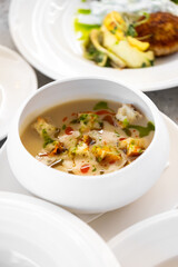 A creamy soup served in a white bowl, garnished with croutons, fresh herbs, and oil drizzle. Background shows a plate of vegetables and patty. Perfect for culinary and food photography themes.