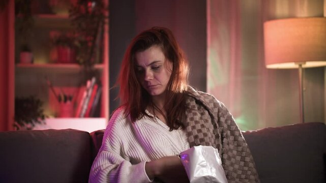 Cute red-haired woman sitting on sofa, shaking off food remains, looking around tiredly after watching TV
