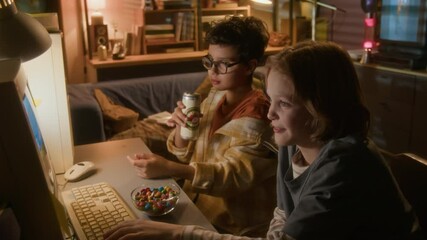 High angle shot of two young boys playing videogame and eating candy in nostalgic 90s setting, focus on boy wearing glasses and pointing at computer screen helping friend
