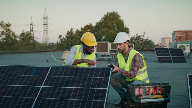 Two multiethnic engineers collaborate on solar panel installation, examining connections and ensuring efficiency. A toolbox with essential equipment is nearby on the rooftop. Males workers on workday.