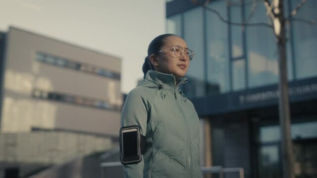 Female jogger, a young Asian woman wearing active sportswear, setting her app on her phone. Soft sunlight. Health and outdoor activity. Sports tech.