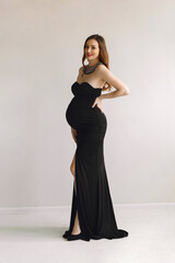 Happy young pregnant woman in stylish black dress and ethnic necklace posing on light wall. Stylish...
