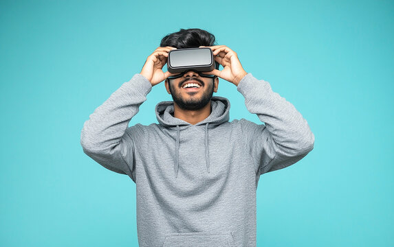 Excited young Indian man in a gray hoodie holding a VR headset near his eyes, captured in a studio shot against a pastel blue background. Perfect for technology, gaming, and virtual reality concepts.

