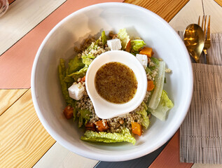 Fresh quinoa salad served in white bowl with lettuce, feta cheese, roasted pumpkin, nuts, flavorful dressing in separate dish on wooden table , healthy gluten free plant based breakfast option