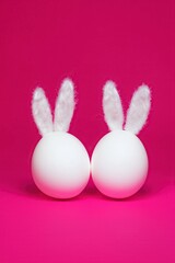 Two white eggs with fluffy white bunny ears placed side by side against vibrant pink backdrop