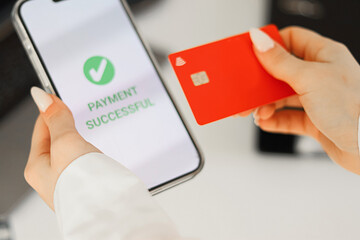 Hand holding a red bank card and a smartphone with a successful payment message on the screen. A laptop is visible in the background. Secure online payment, remote shopping, and digital transaction.